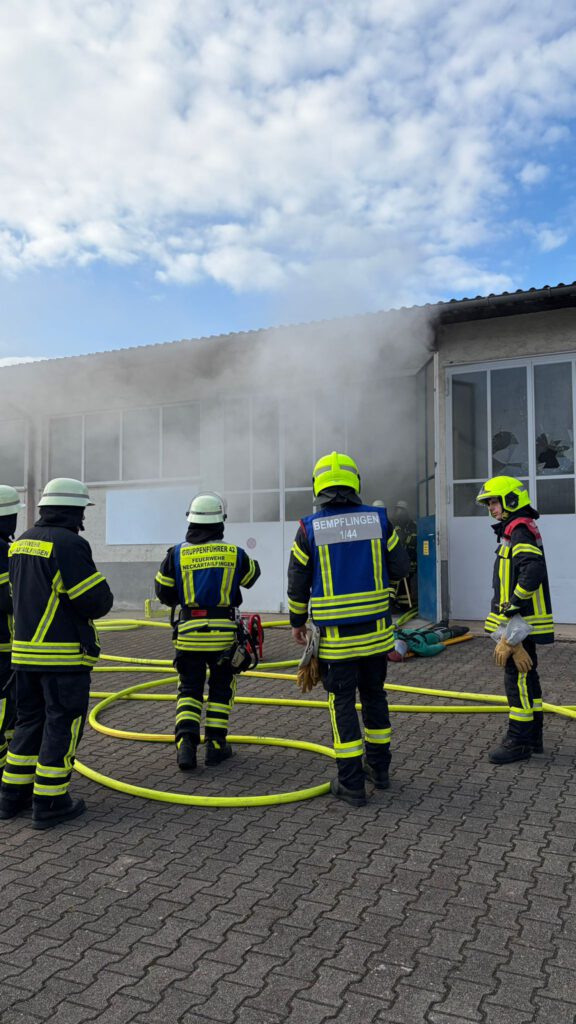 Die FF Bempflingen und die FF Neckartailfingen bekämpfen einen Brand in einer Lagerhalle.
Auf dem Bild sind mehrere Personen der Feuerwehr zu sehen, welche vor einer offenen Hallentüre stehen aus der dichter Rauch rauskommt.