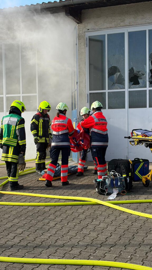 Die Malteser nehmen die verletzten Personen entgegen.
Auf dem Bild ist zu sehen, wie die Feuerwehr eine verletzte Person auf der verrauchten Lagerhalle an die Rettungskräfte der Malteser übergibt.